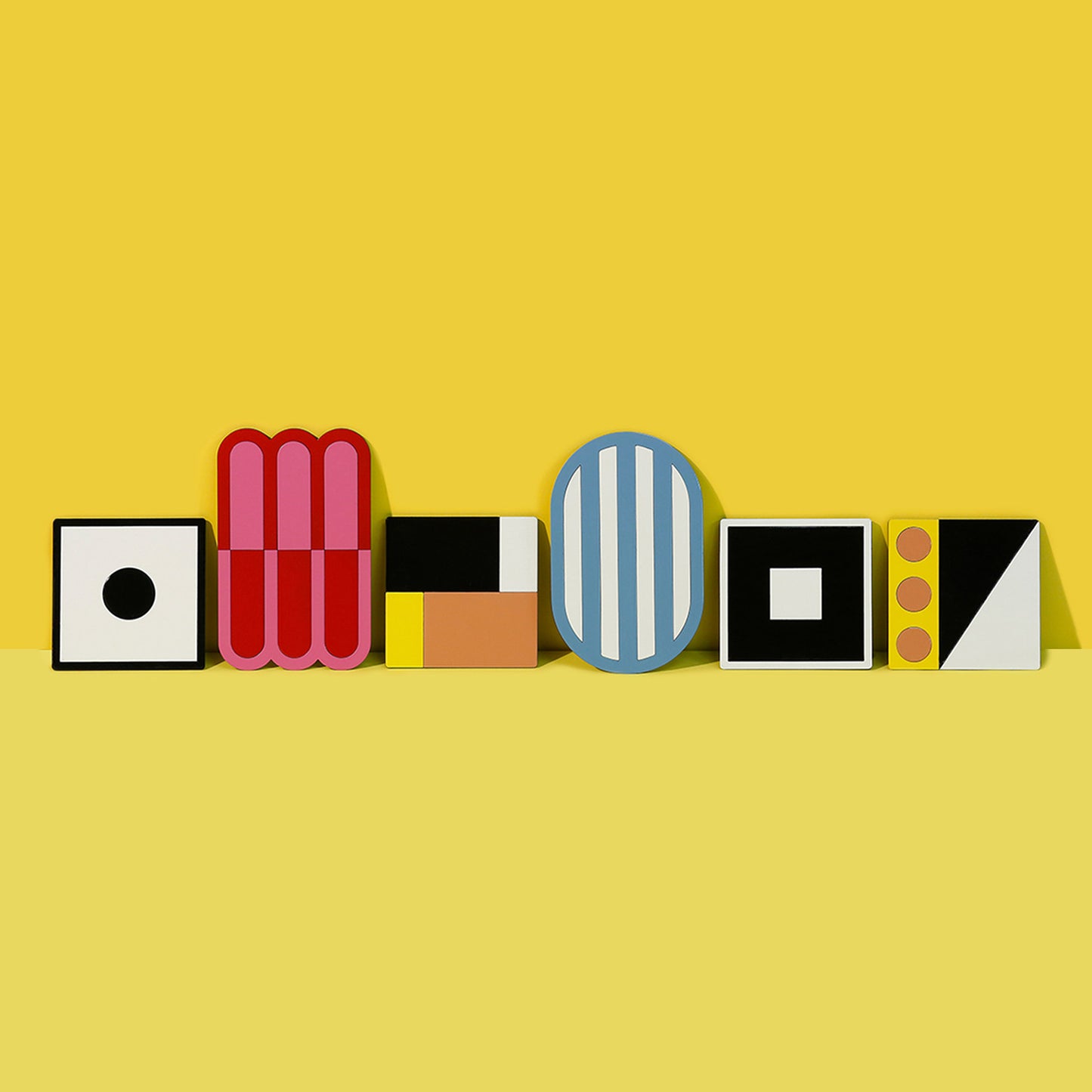 A lineup of six bold and artistic metal coasters in various geometric shapes and colors, displayed upright against a bright yellow background – ISUWA Coaster Collection.