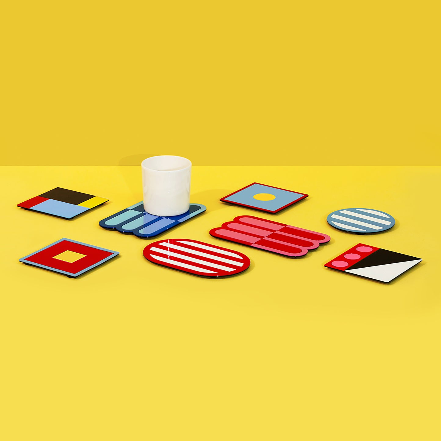 Colorful assortment of geometric metal coasters including round, square, and wave-shaped designs with one white cup placed on a blue wave coaster – vibrant display on yellow surface