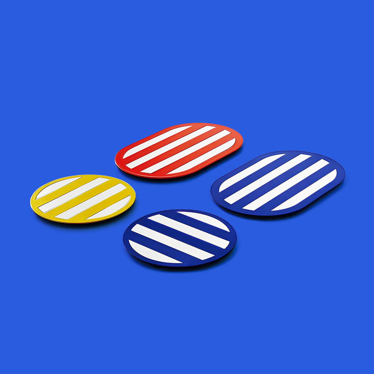 Four striped metal coasters in vibrant red, yellow and navy blue, with white diagonal lines — shown on a bold blue background, modern and graphic ISUWA design.