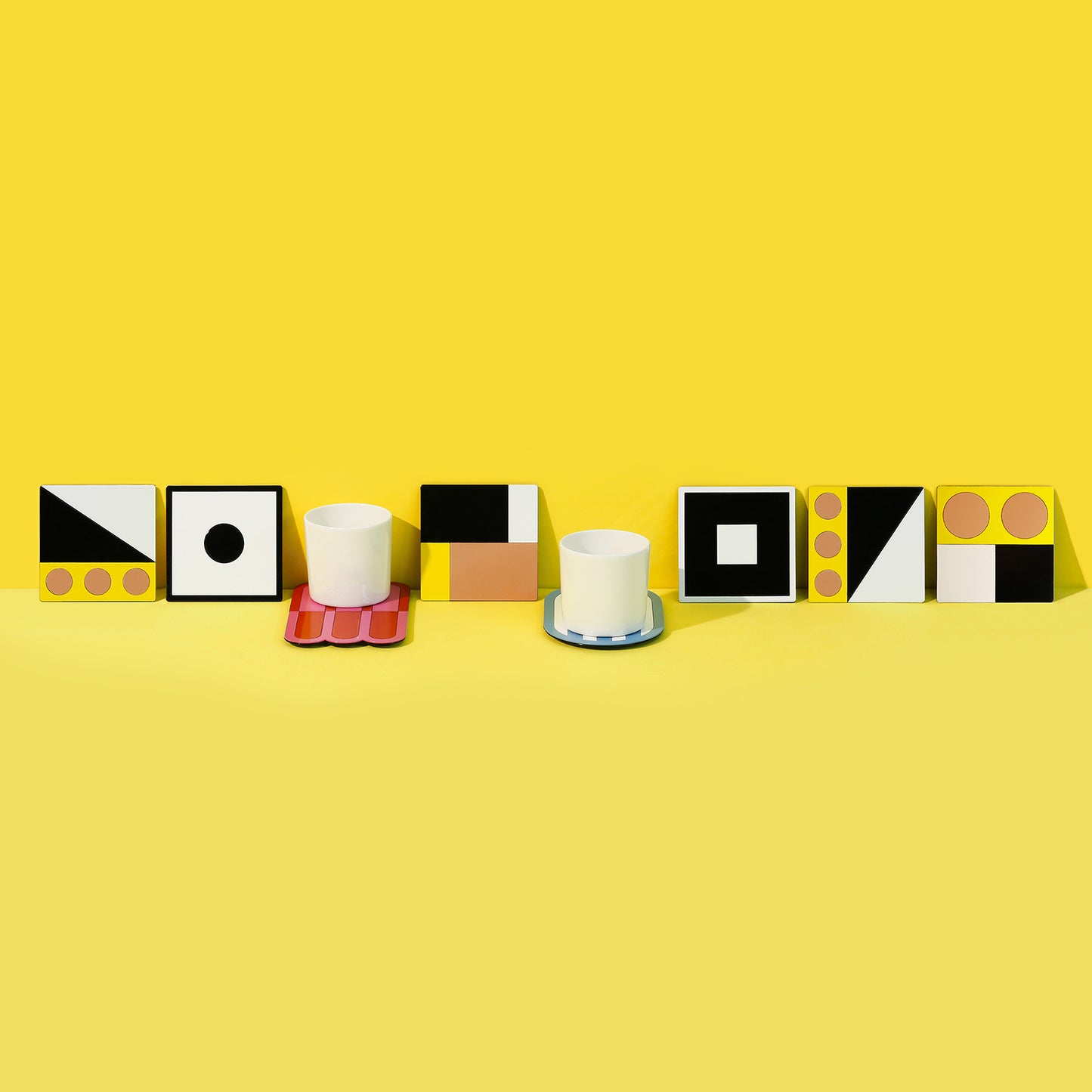 A row of white porcelain cups placed on and behind glossy geometric coasters in bold colors and shapes, including circles, squares and diagonals. Set against a vibrant yellow background, the composition highlights ISUWA’s playful and artistic tableware design.