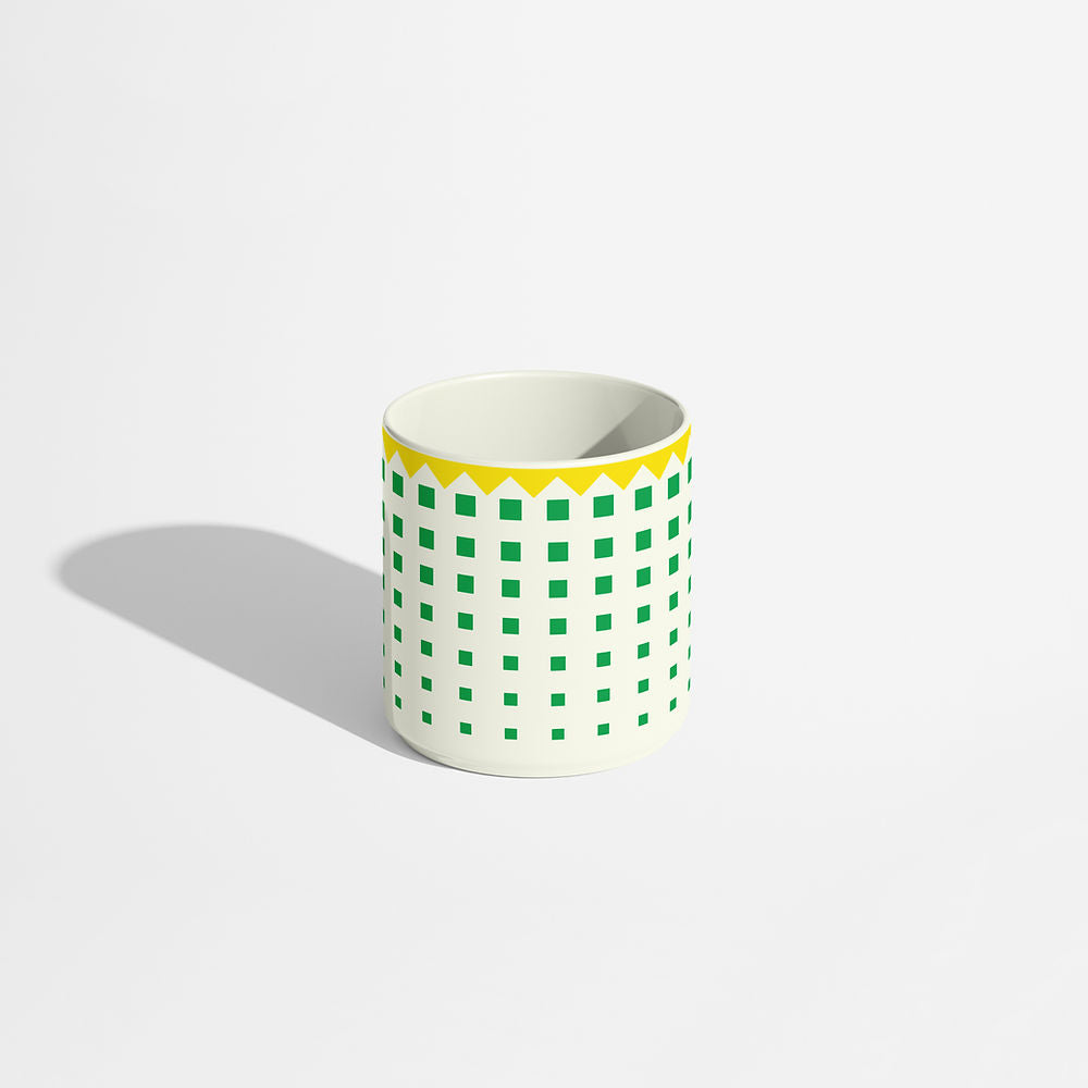 Porcelain coffee cup with a green square grid pattern and yellow zigzag rim, photographed on a light background.
