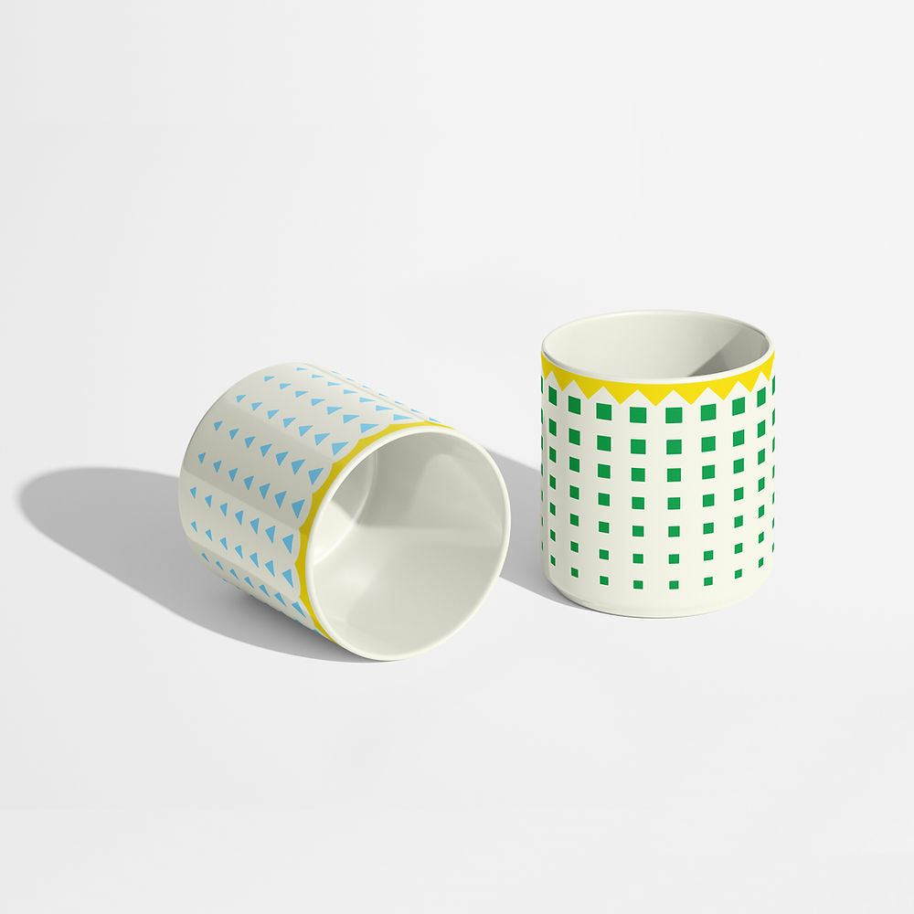 Two porcelain coffee cups with geometric patterns — one with green squares and a yellow zigzag rim, the other with blue triangles — styled on a light surface.
