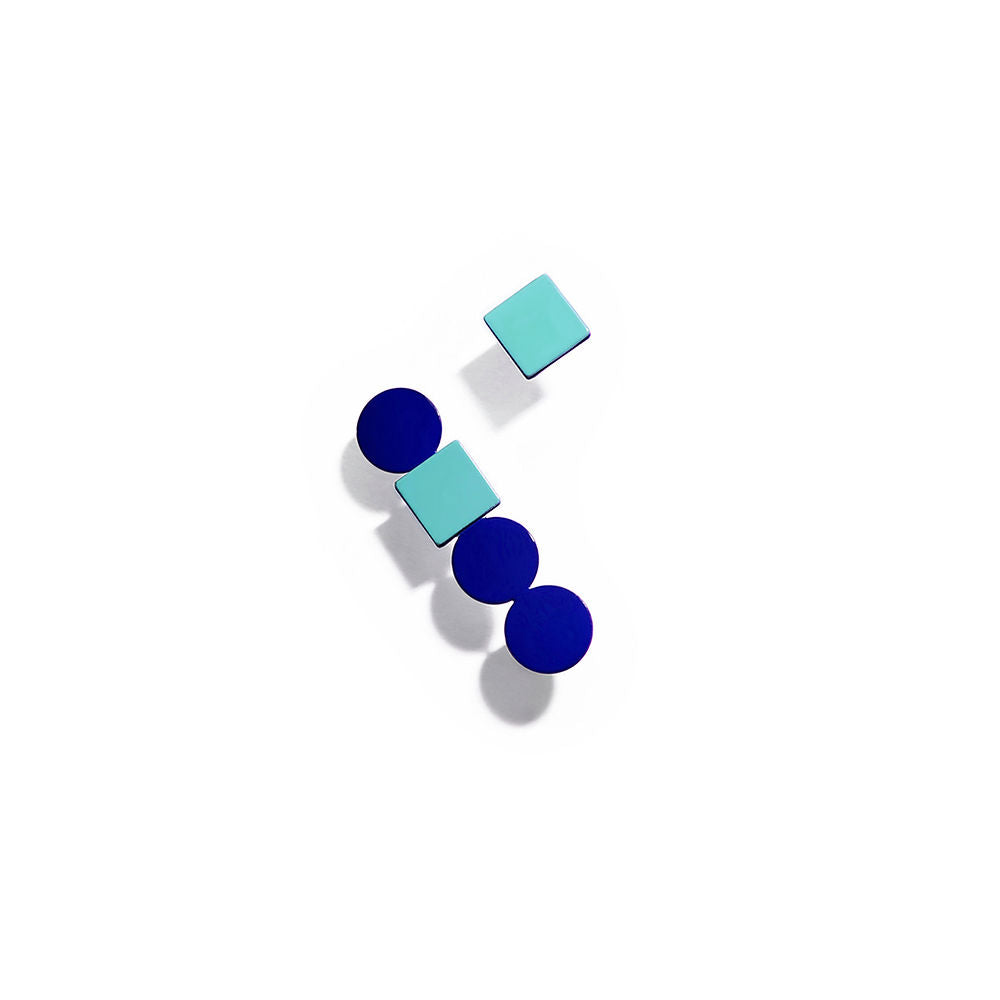 A pair of asymmetrical earrings combining geometric shapes in vibrant colors. One earring features two blue circles and a mint green square, while the other has a mint green square and a blue circle. Bold, modern composition on a white background with strong shadows.
