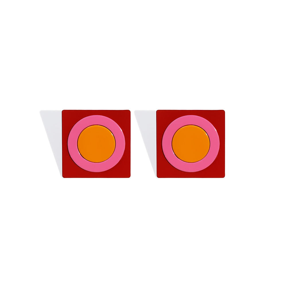 A pair of square stud earrings featuring bold concentric shapes: an orange circle at the center, surrounded by a pink ring, all set on a red square base. Photographed on a white background with strong shadow contrast.
