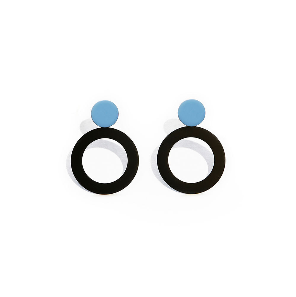 A pair of modern geometric earrings featuring a small solid blue circle on top of a large black open circle. Minimalist and bold design, photographed on a white background with soft shadows.