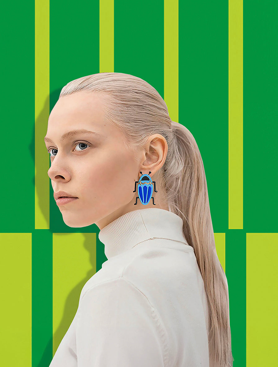 Woman wearing sculptural blue bug earrings by ISUWA, handmade jewelry with bold and modern design on green patterned background