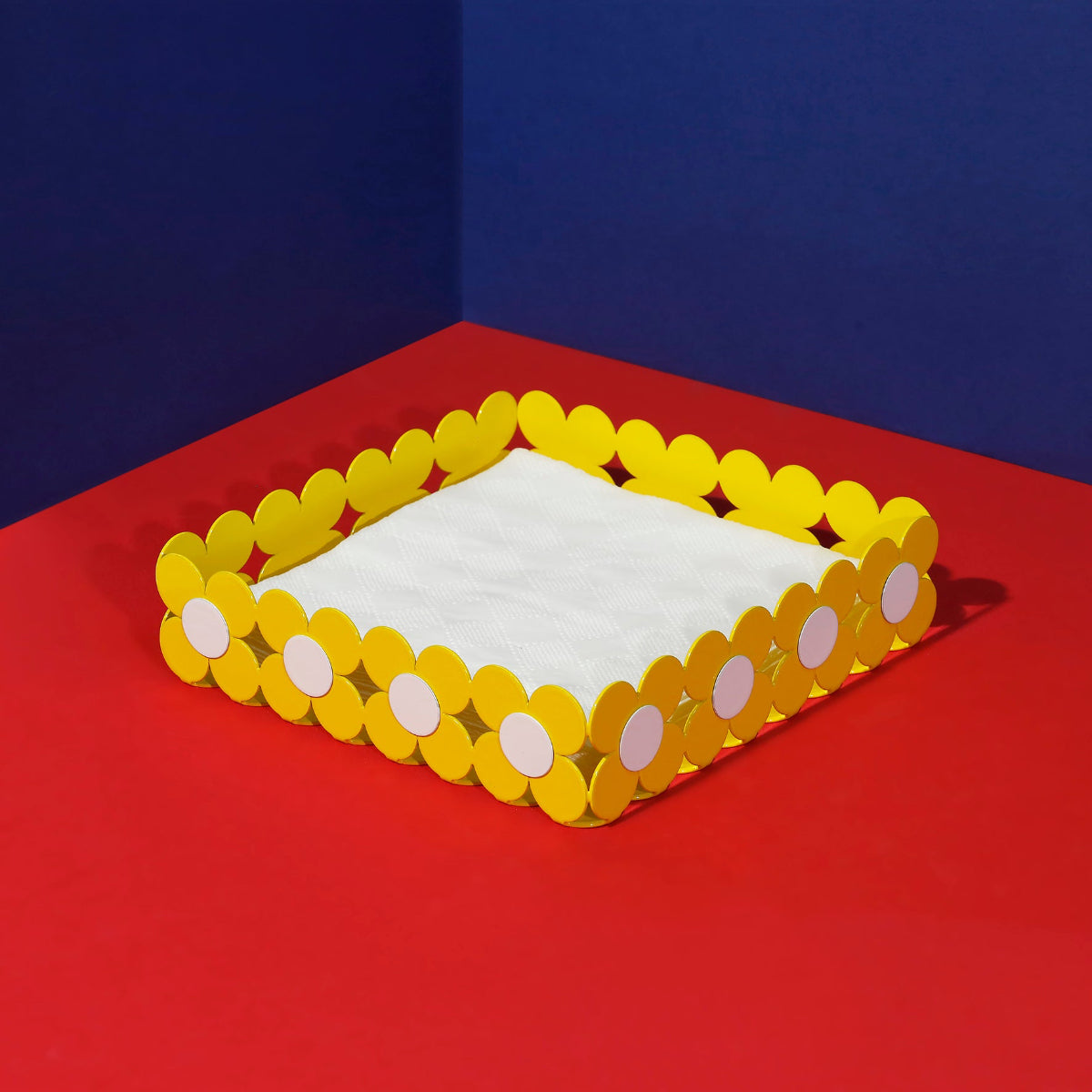 ISUWA yellow flower-shaped metal napkin holder on a red surface with a blue backdrop, holding white napkins.