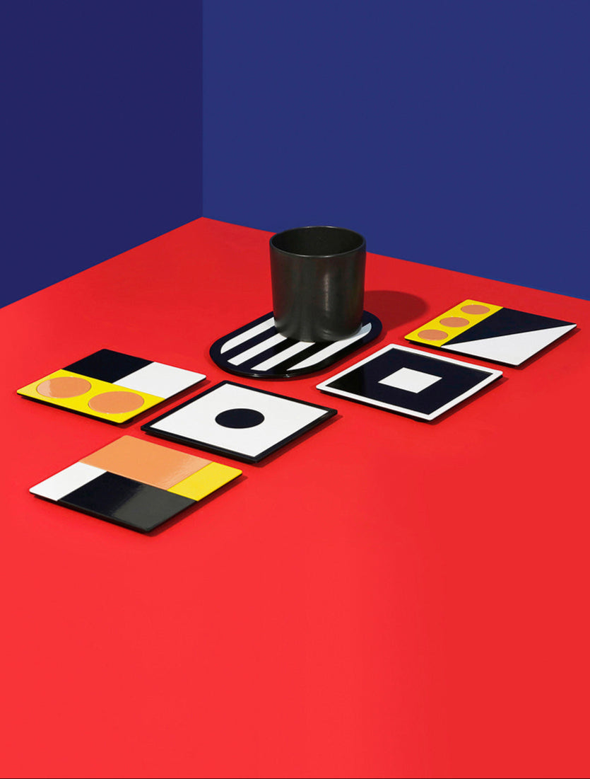ISUWA Geometric Coasters – artistic metal coasters in bold graphic patterns and colors including yellow, black, white and peach, displayed on a red surface with a blue wall background.