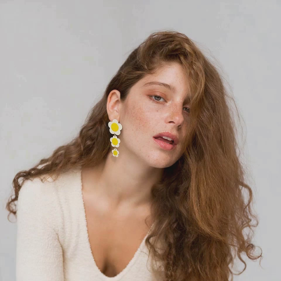 Model with natural makeup and loose auburn hair showcasing yellow daisy statement earrings in a calm studio setting, emphasizing the earrings' playful design and vibrant colors