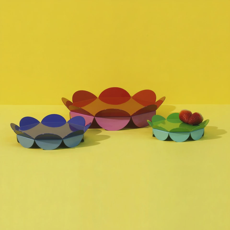 Colorful flower-shaped ISUWA trays in blue, red-pink, and green-aqua displayed on a vibrant yellow surface. The smallest tray holds a few bright red strawberries. Modern, joyful, and sculptural.