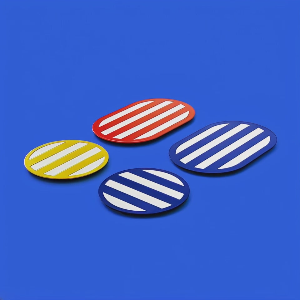 Four glossy metal coasters with diagonal white stripes in red, yellow, navy blue, and royal blue. Arranged on a vibrant blue background, showcasing their oval and round shapes in a bold, geometric composition.