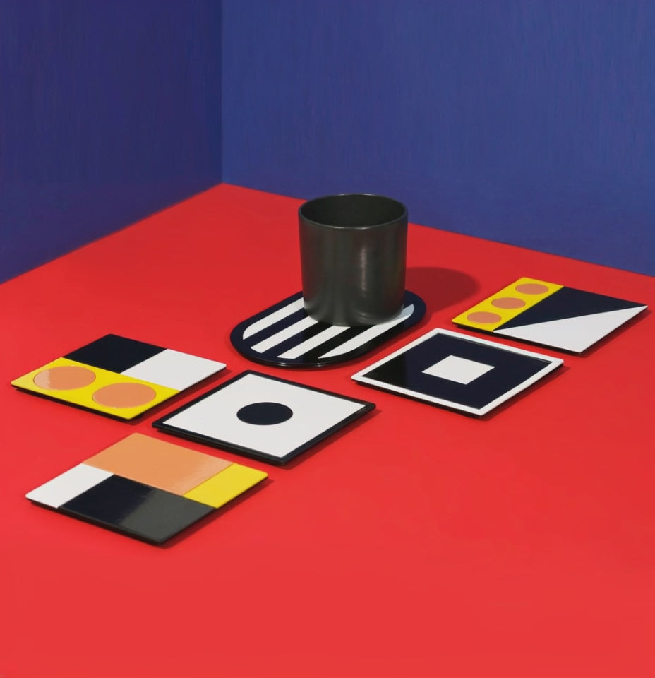 Video showing a black porcelain cup on a black striped oval coaster surrounded by various colorful geometric square coasters – an artistic and playful setup on red and blue background.