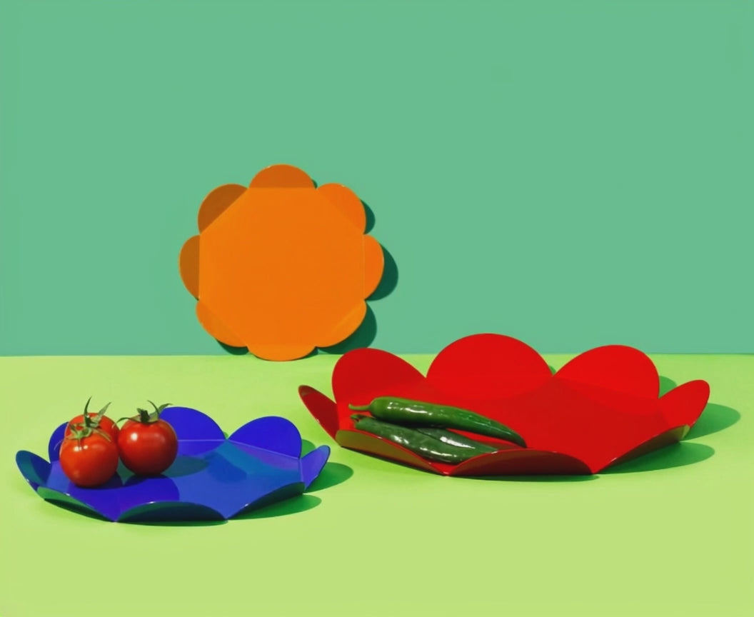 A rotating view of three flower-shaped metal trays in navy, red and orange. The navy tray holds tomatoes, the red tray holds green peppers, and the orange tray is placed upright in the background. Set against a green surface and backdrop.
