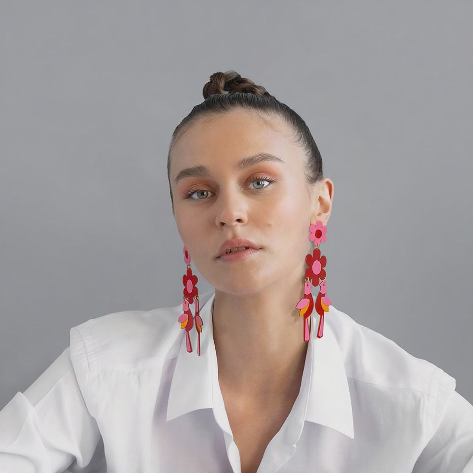Model wearing bold red bird statement earrings with pink flower tops and orange accents, styled with a white shirt against a neutral background – showcasing the earrings’ length and sculptural movement.
