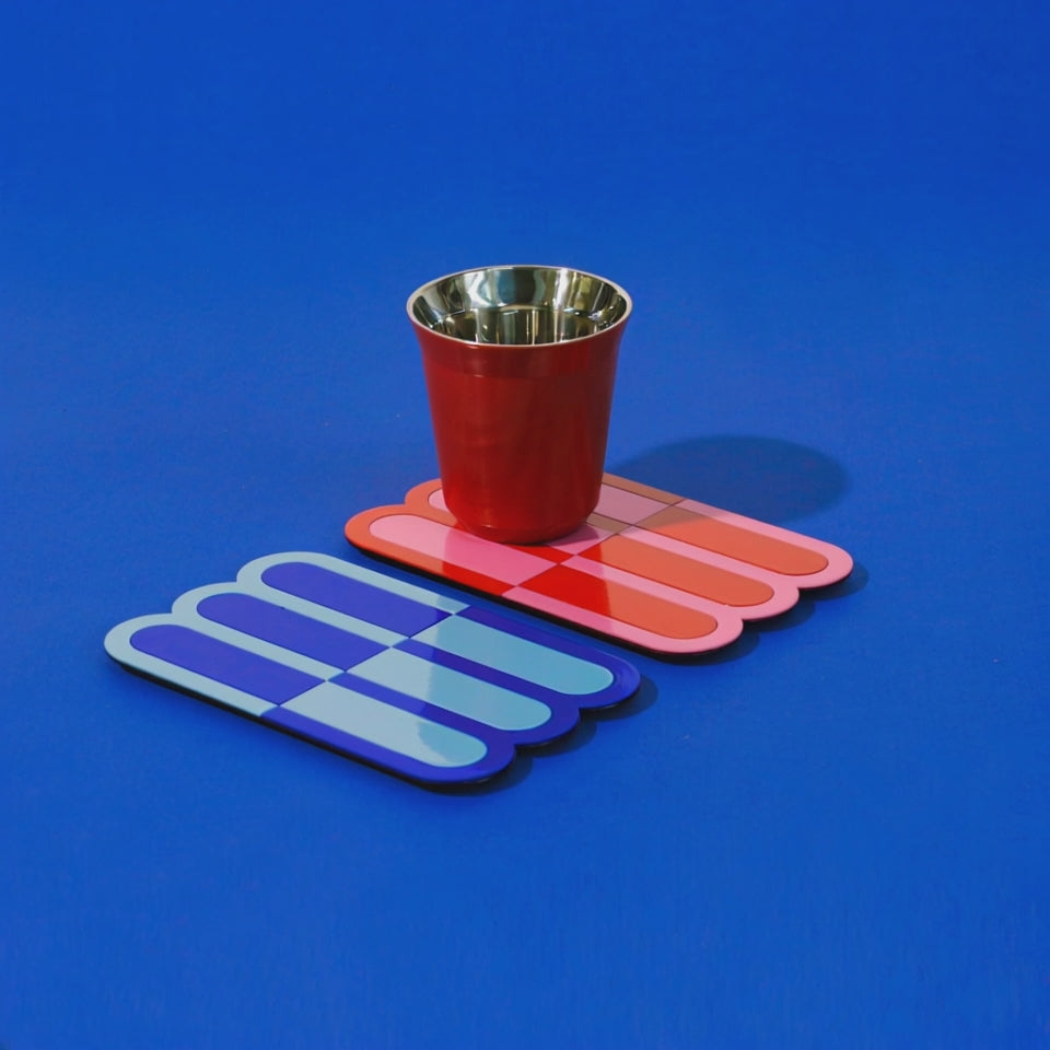 A video showing a red metal cup placed on a red wave coaster, beside a blue wave coaster, all set against a striking blue background – artistic tabletop arrangement.