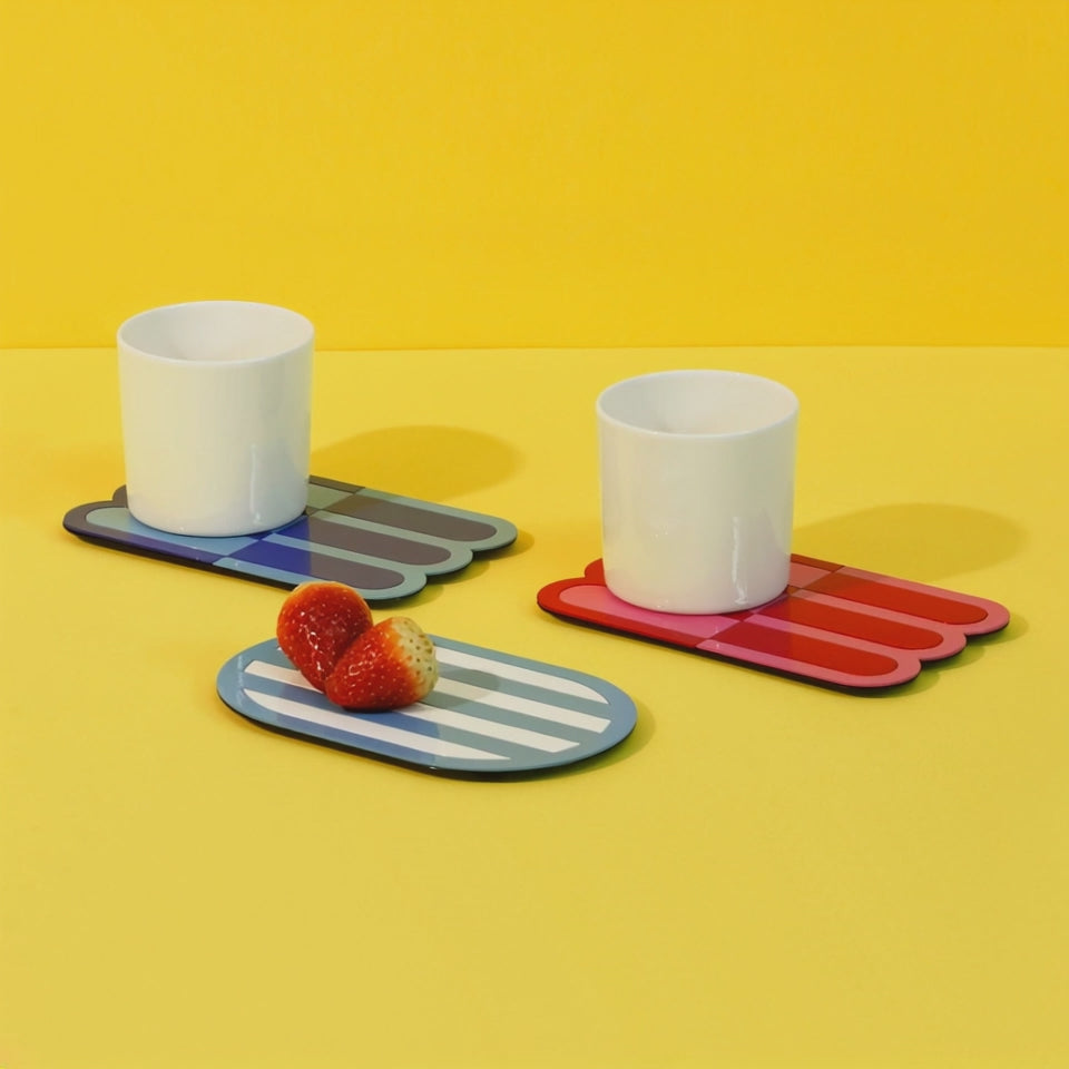 Video showing two white cups placed on red and blue wave-patterned metal coasters, with strawberries on a striped oval coaster – artistic table setup on a bright yellow background.